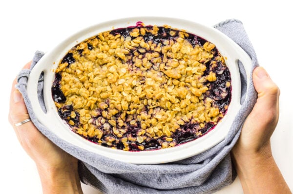 Hands hold a towel around a freshly baked batch of blueberry crumble.