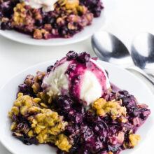Two plates full of blueberry crumble with ice cream on top. There are two spoons between them.