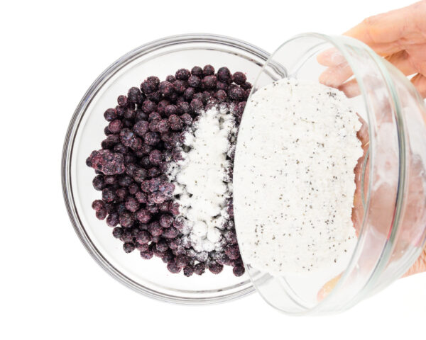Two hands hold a bowl of flour mixture, pouring it in with a bowl of frozen blueberries.