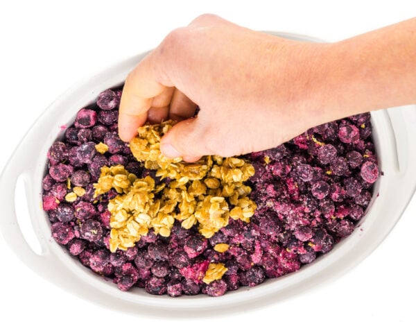 A hand holds crumble mixture and is spreading it across blueberries in a casserole dish.