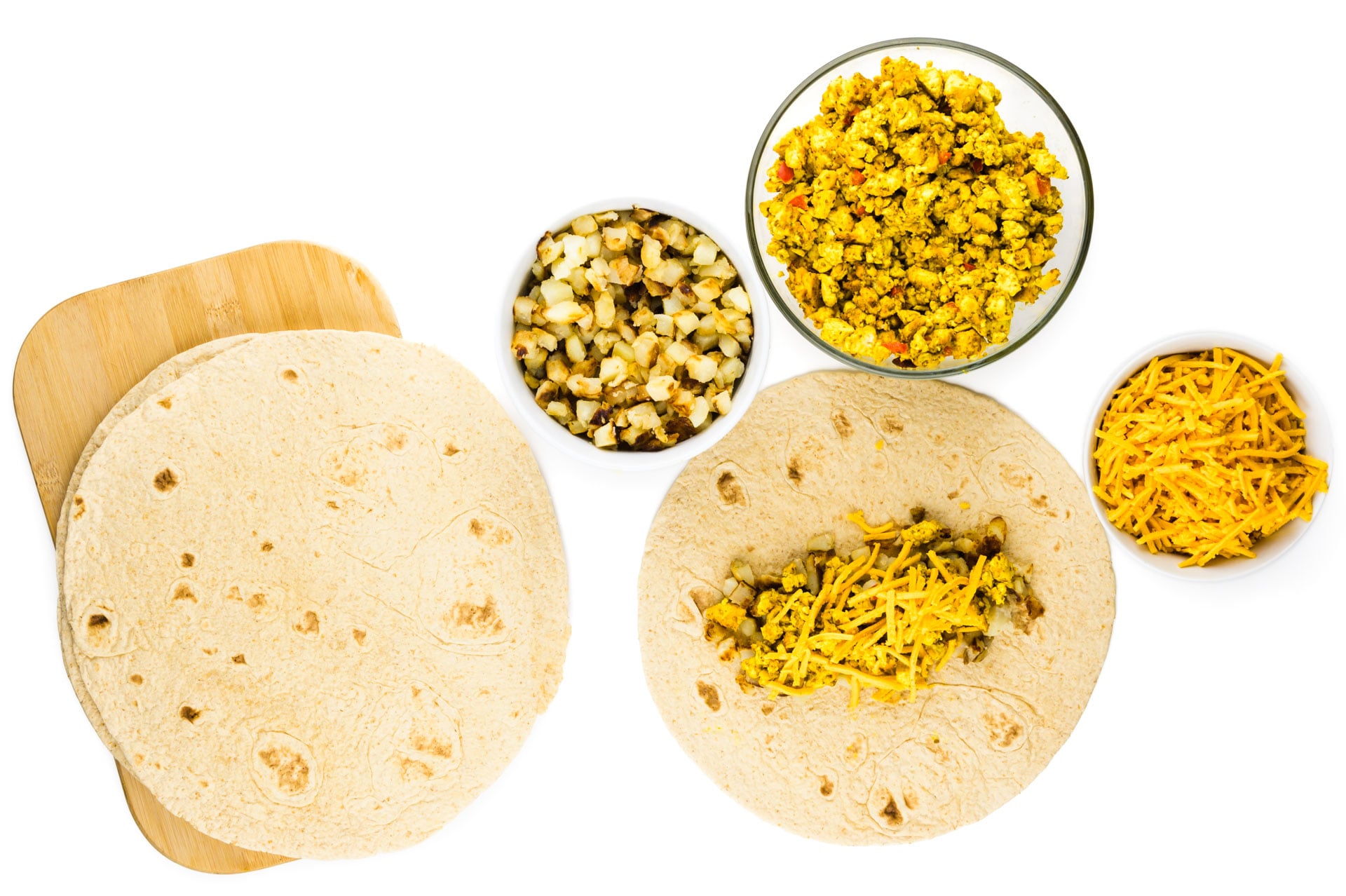 A tortilla holds tofu scramble and other ingredients in bowls around it to make breakfast burritos.