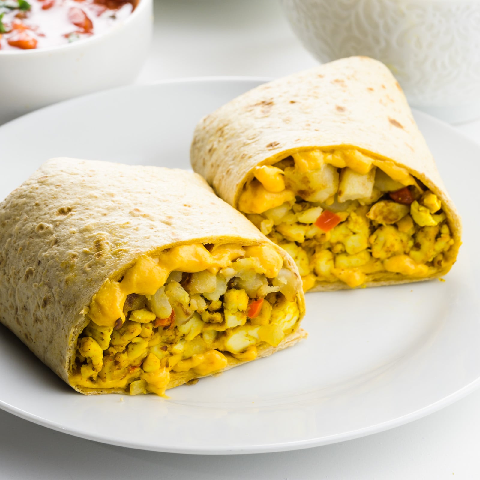 Two halves of a breakfast burrito on a plate with a bowl of salsa behind it.