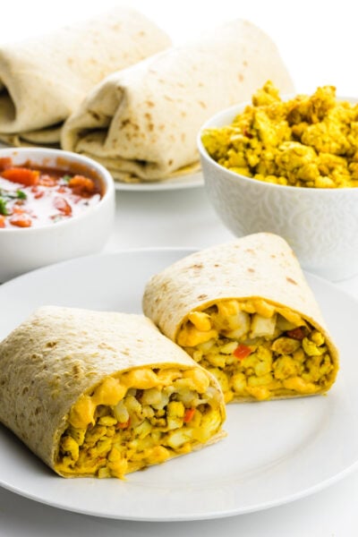 A vegan breakfast burrito has been cut in half showing the cheesy insides. A bowl of salsa is behind it, along with a bowl of scrambled tofu and more burritos.