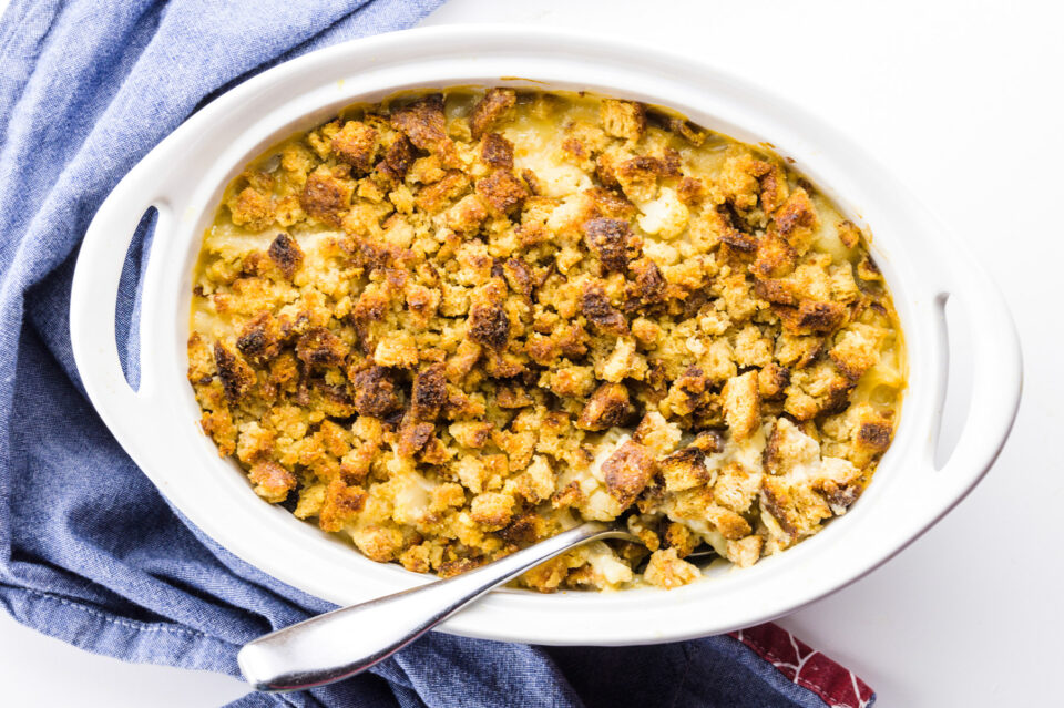Looking down on a casserole dish with bread crumbs on top. There's a spoon in the dish and a blue kitchen towel on the left.