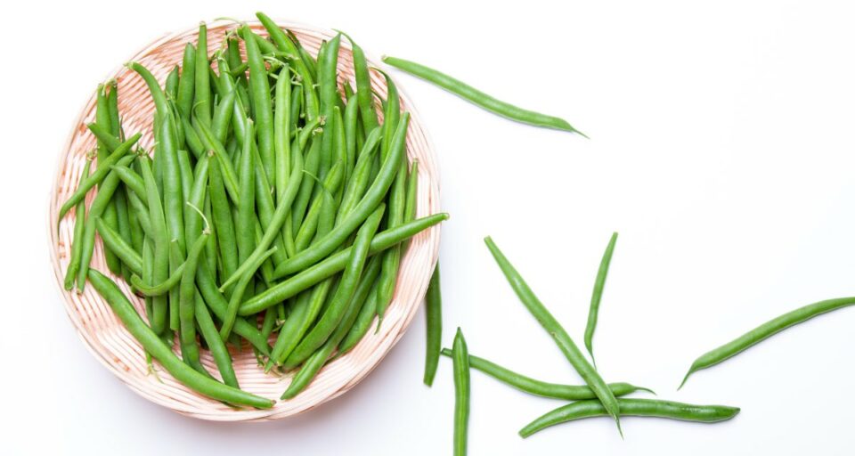 Looking down on a basket full of fresh green beans. There are green beans on the table next to the basket, too.