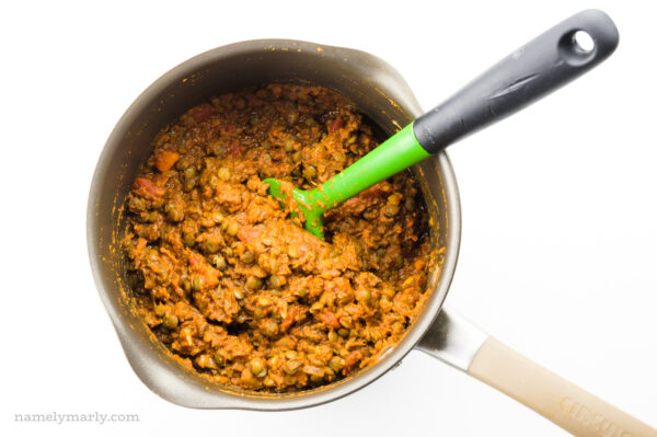 Looking down on a saucepan full of lentils and veggies in a tomato sauce. A green spatula is in the pot for stirring.