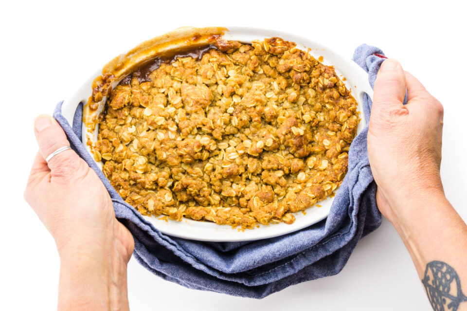 Two hands hold a blue kitchen towel at both ends of a casserole dish full of an apple dessert.
