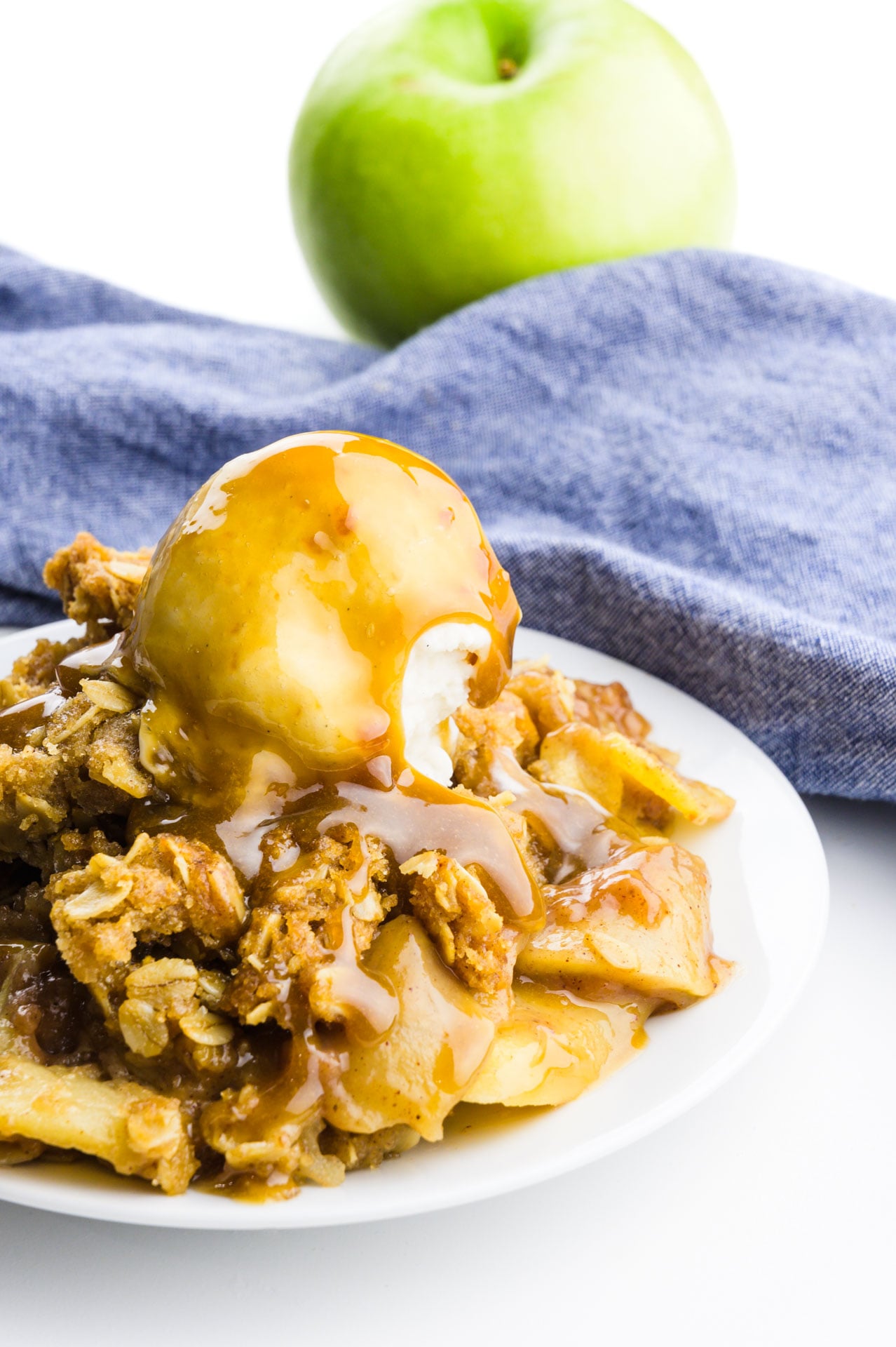A plate holds an apple dessert with ice cream on top and caramel sauce drizzled over the top. A blue towel and green apple are behind it.
