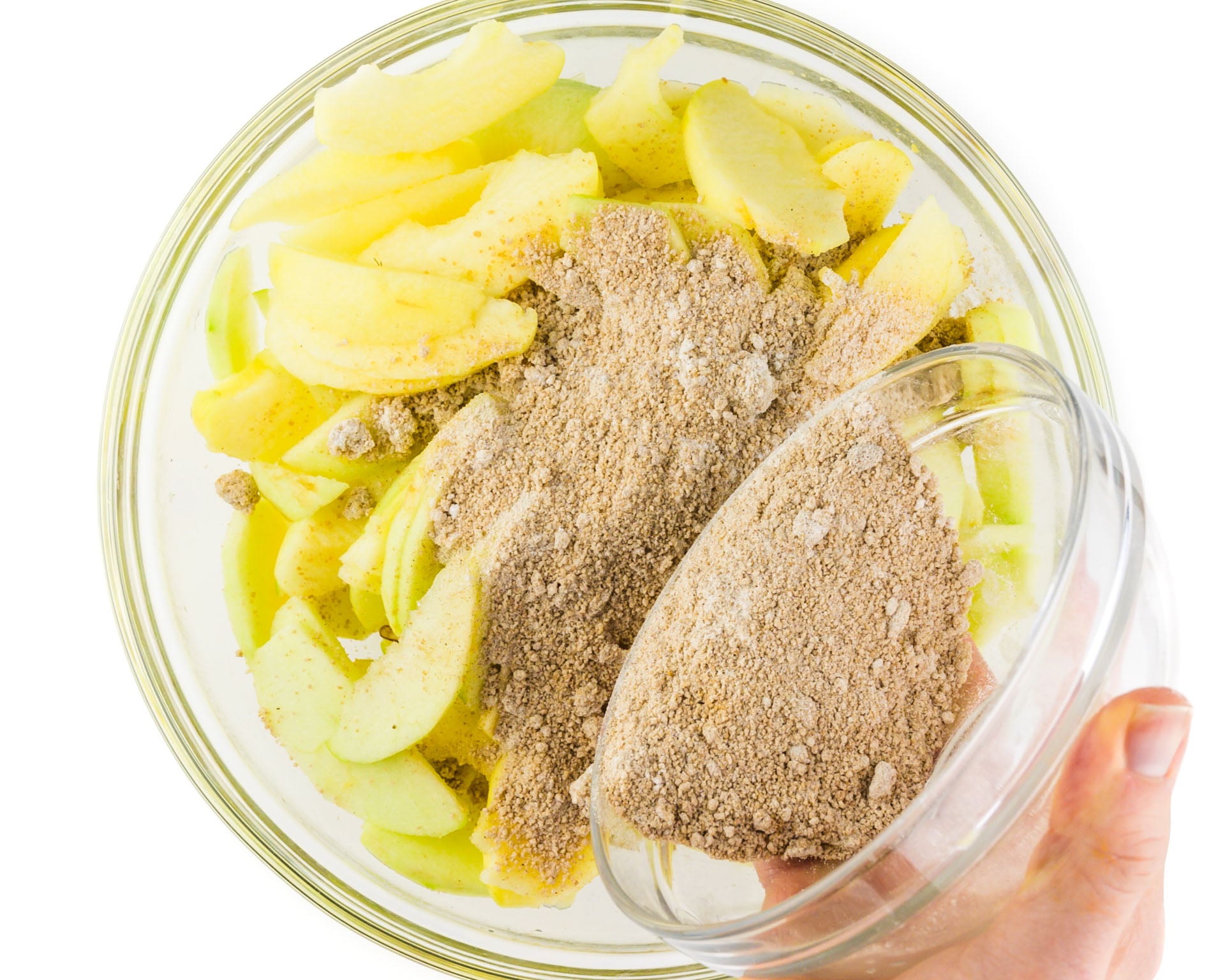 A hand holds a bowl pouring cinnamon mixture over sliced apples in a bowl.