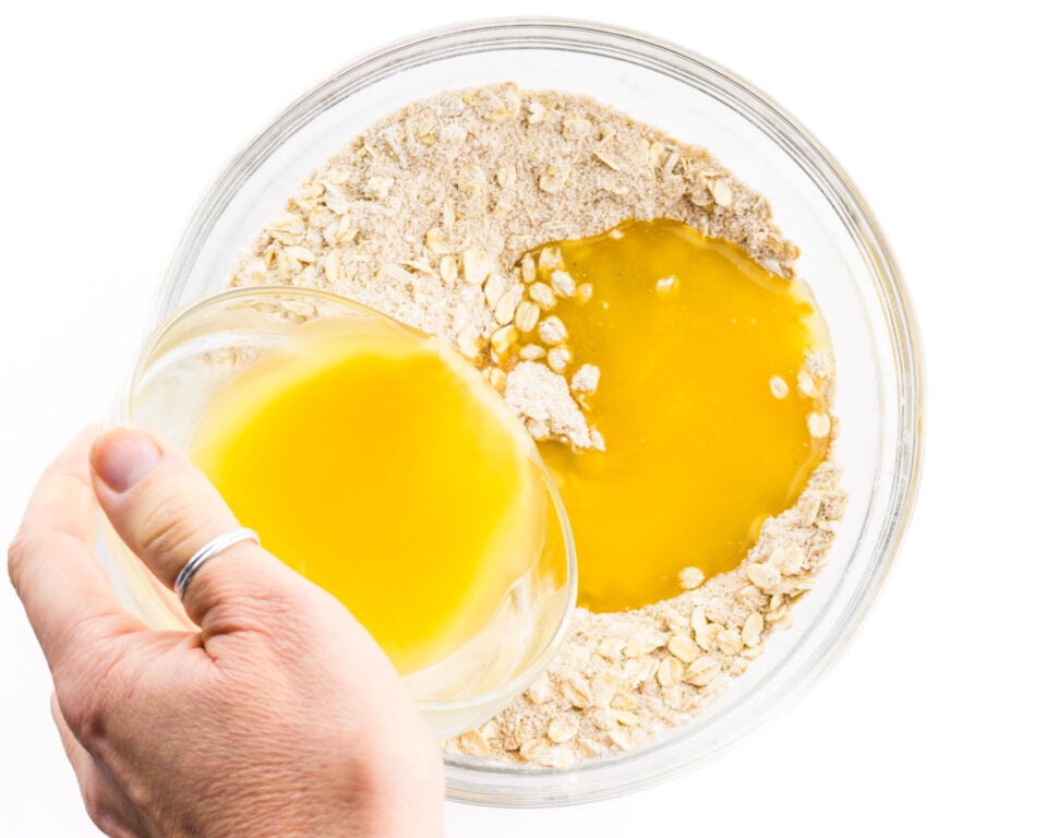 A hand holds a bowl of melted butter and is pouring it over a bowl full of a flour mixture.