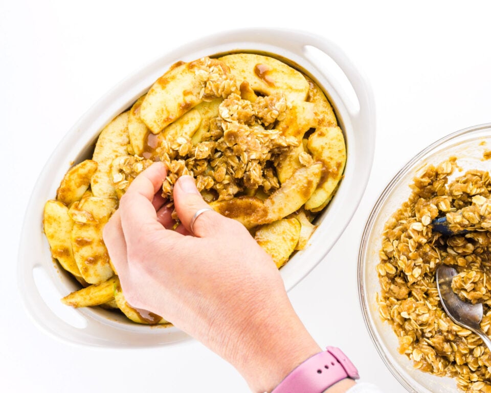 A hand distributes crumble mixture over sliced apples in a casserole dish.