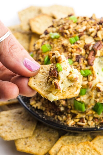 A hand holds a cracker with a bite of vegan cheese ball on it. The cheese ball is behind it with more crackers around it.