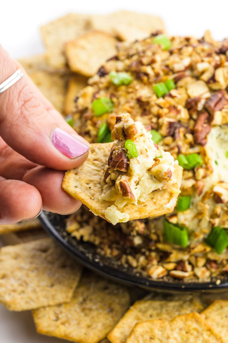 A hand holds a cracker with a bite of vegan cheese ball on it. The cheese ball is behind it with more crackers around it.