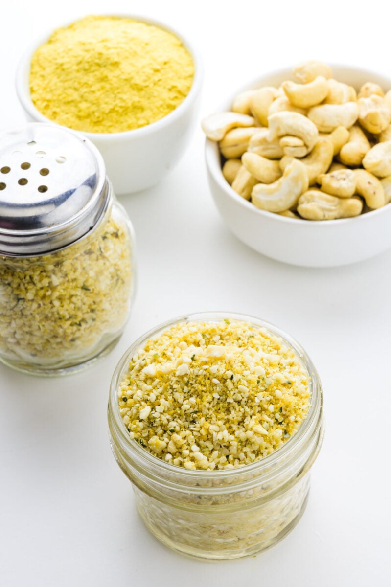 A jar of vegan parmesan sits in front of a bowl with raw cashews, nutritional yeast flakes, and a shaker full of more parm.