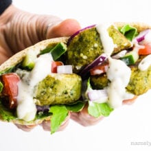 A hand holds a baked falafel sandwich with greens and sauce.