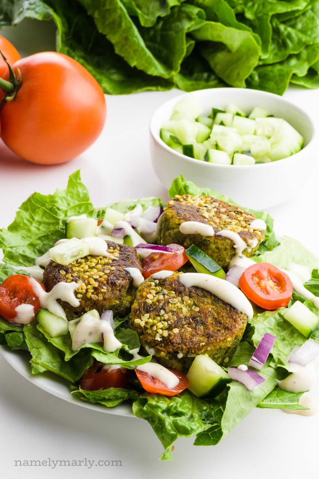 A salad has tomatoes, cucumber, and falafel on top. There is more lettuce, tomatoes, and sauce in the background.