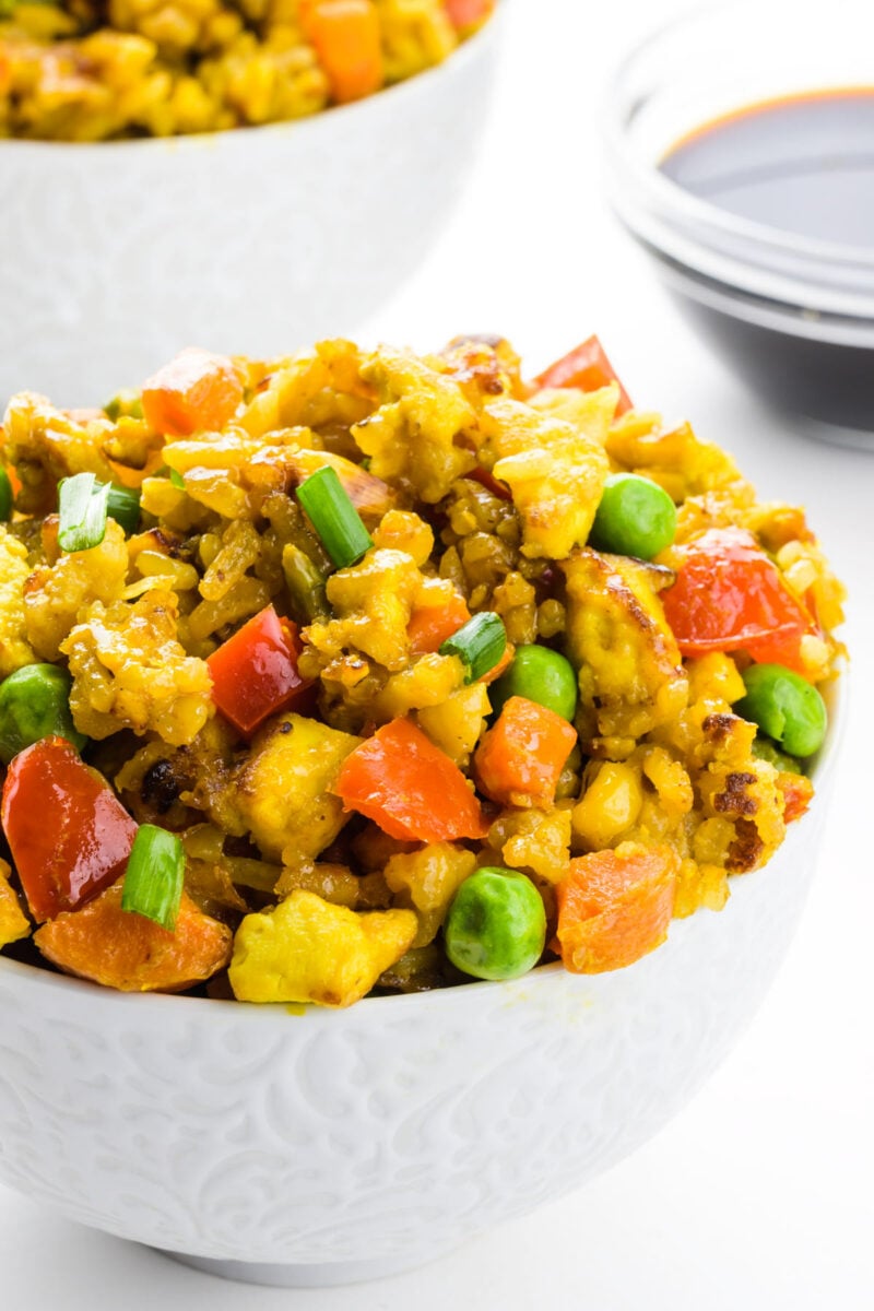 A bowl full of vegan fried rice sits in front of another bowl and a dish of soy sauce.
