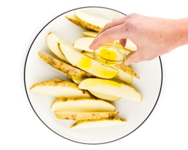 A hand holds a small bowl of oil, drizzling it over cut potatoes.