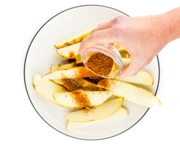 A hand holds a small bowl of spices, pouring it over potatoes wedges on a plate.