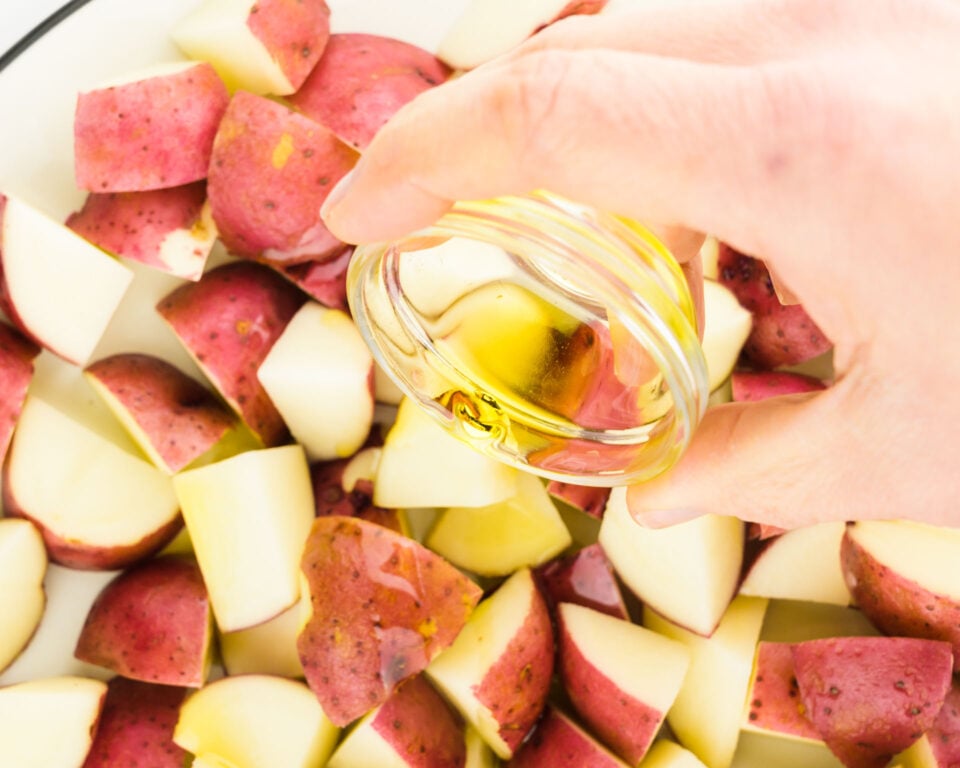 A hand holds a small bowl of oil and is drizzling it over cut potatoes.
