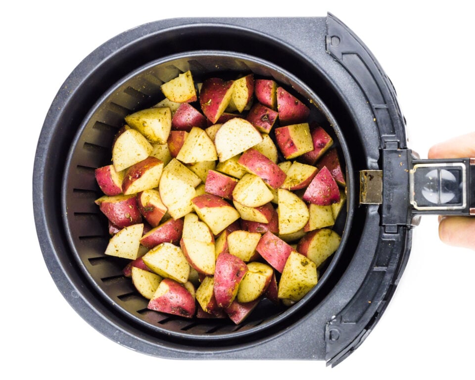A hand holds an air fryer basket full of cut, seasoned potatoes.