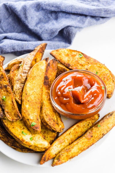 Air fryer potato wedges on a plate with a bowl of ketchup and a blue kitchen towel behind it.