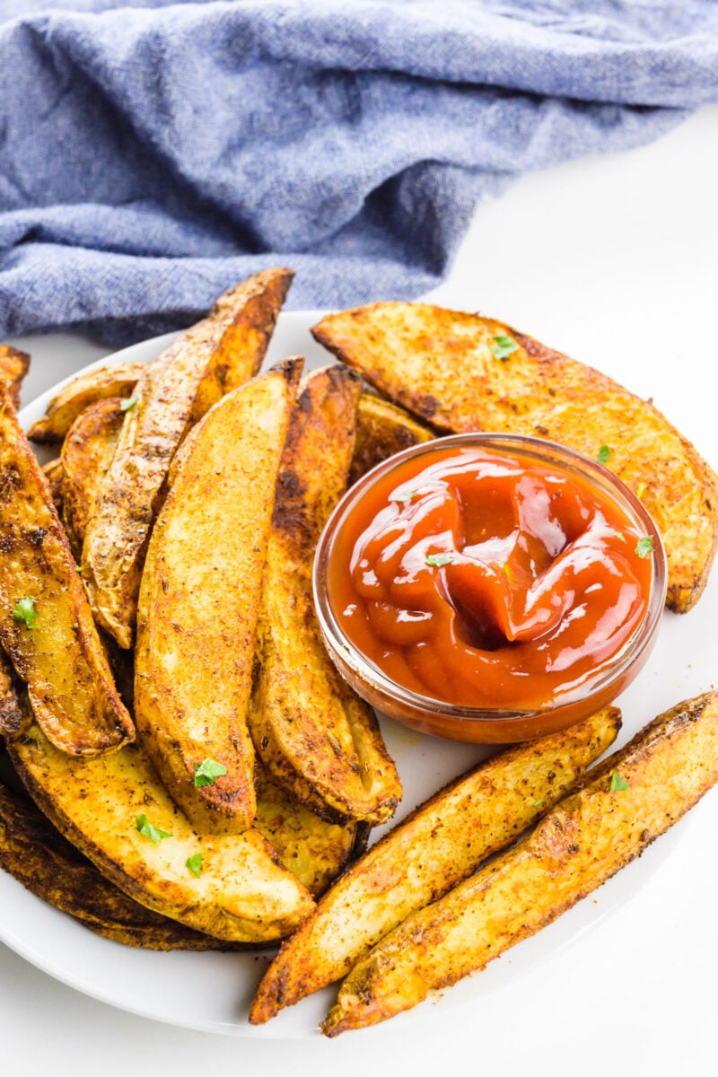 Air fryer potato wedges on a plate with a bowl of ketchup and a blue kitchen towel behind it.