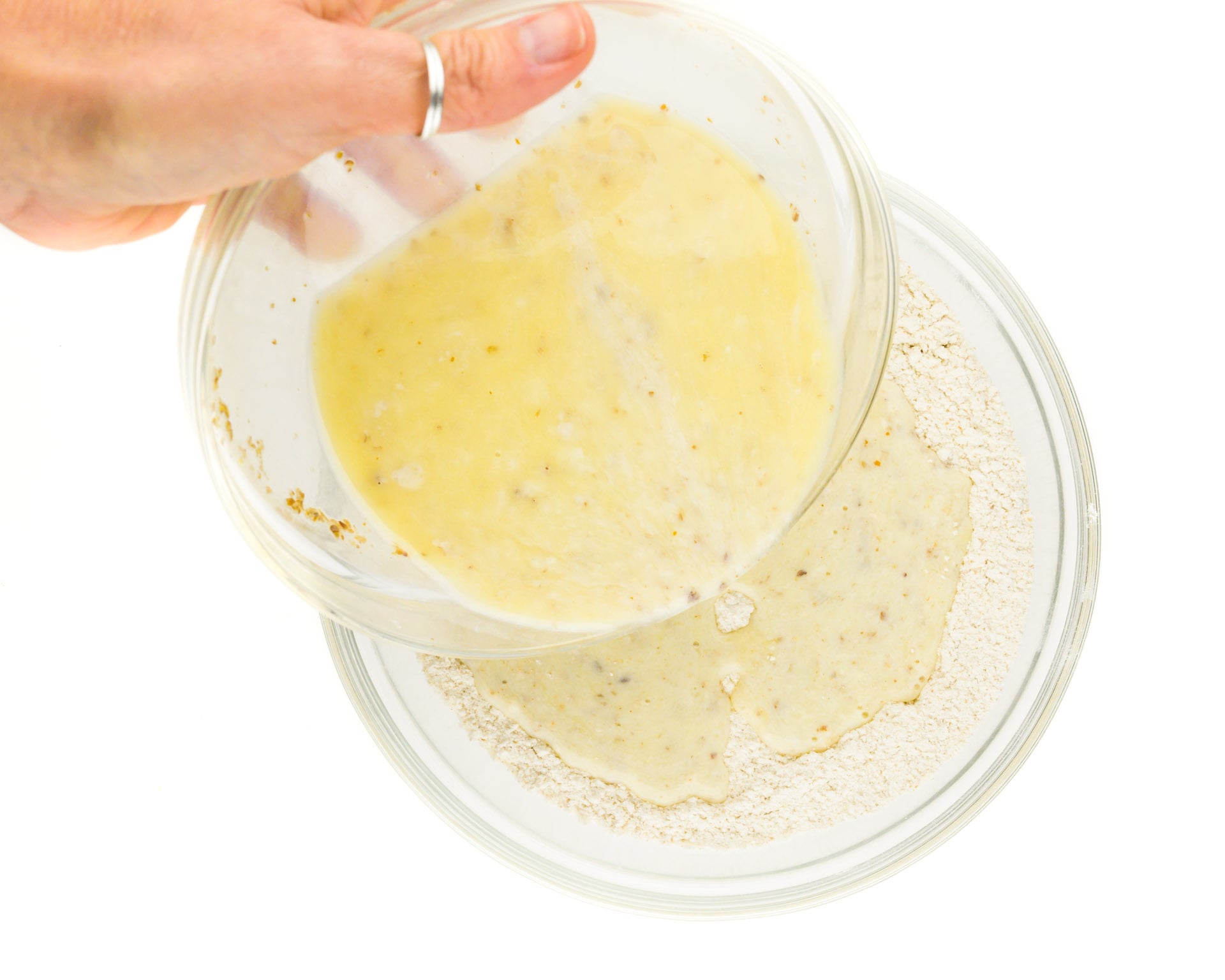 A hand holds a bowl of liquid ingredients and is pouring it into a bowl of flour ingredients.