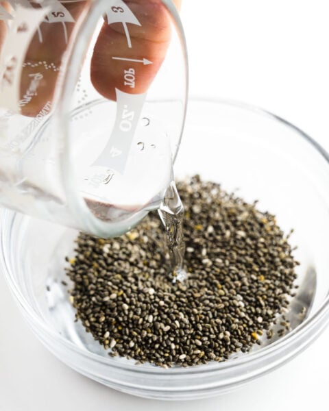 A hand holds a glass measuring cup with water and is pouring it into a bowl with chia seeds.