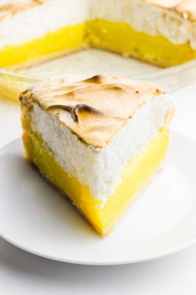 A lemon pie sits on a plate with the rest of the pie behind it.