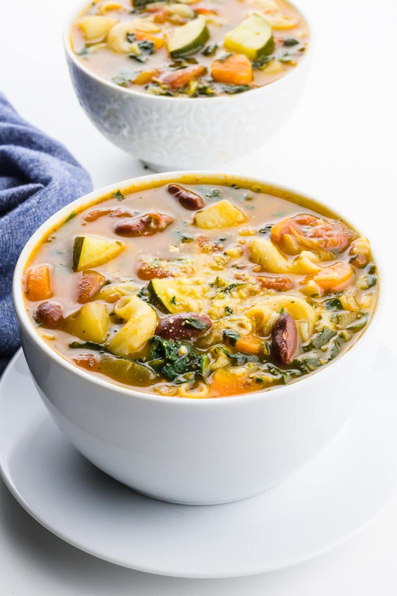 Two Bowls of vegan minestrone are on a table top with a blue kitchen towel between them.