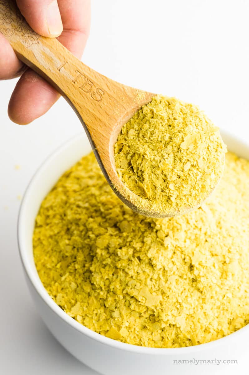 A hand holds a wooden measuring spoon full of nutritional yeast flakes. It's hovering over a bowl full of more of the flakes.