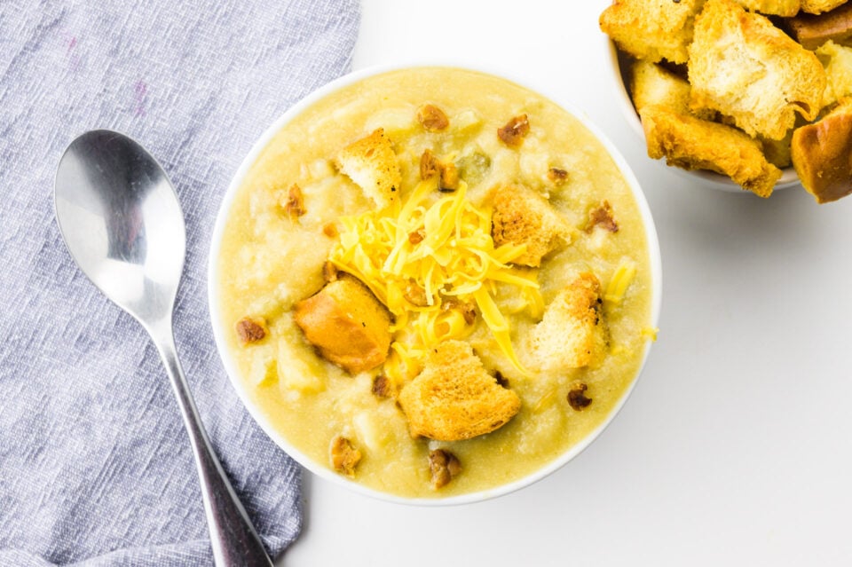 Looking down on a bowl of potato soup with a bowl of croutons on one side and spoon on a grey napkin on the other side.