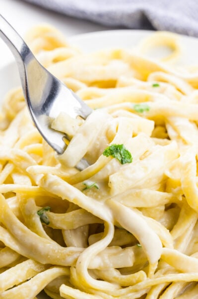 A fork dips into a bowl of fettuccine covered with vegan alfredo sauce and parsley sprigs.