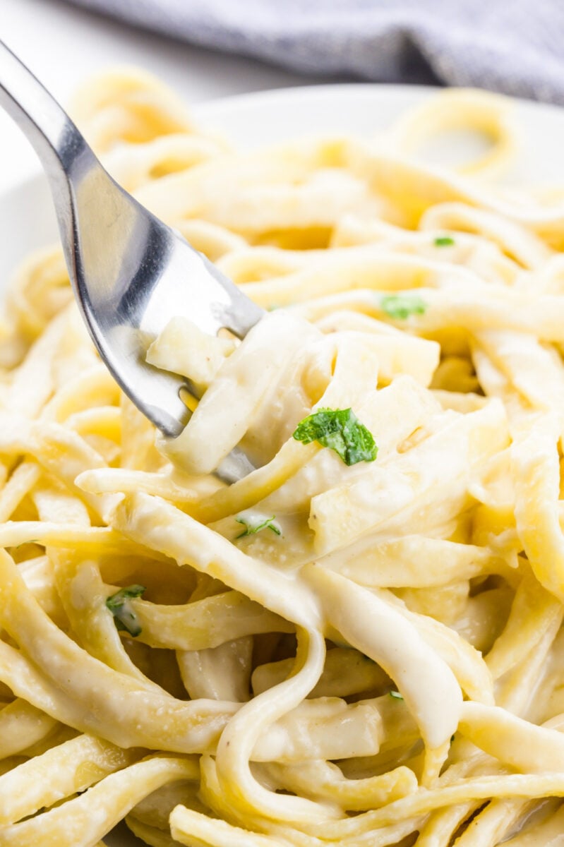 A fork dips into a bowl of fettuccine covered with vegan alfredo sauce and parsley sprigs.
