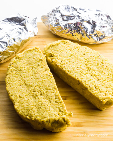 Slices of raw vegan chicken have been cut on a cutting board and more slices wrapped in foil sits behind it.
