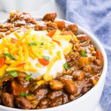 A bowl of vegan chili is topped with sour cream, and vegan cheddar shreds. A bowl of tortilla chips and a blue kitchen towel is behind it.