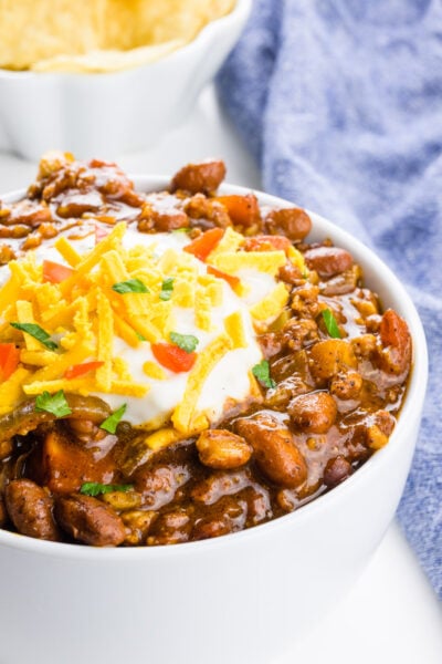 A bowl of vegan chili is topped with sour cream, and vegan cheddar shreds. A bowl of tortilla chips and a blue kitchen towel is behind it.