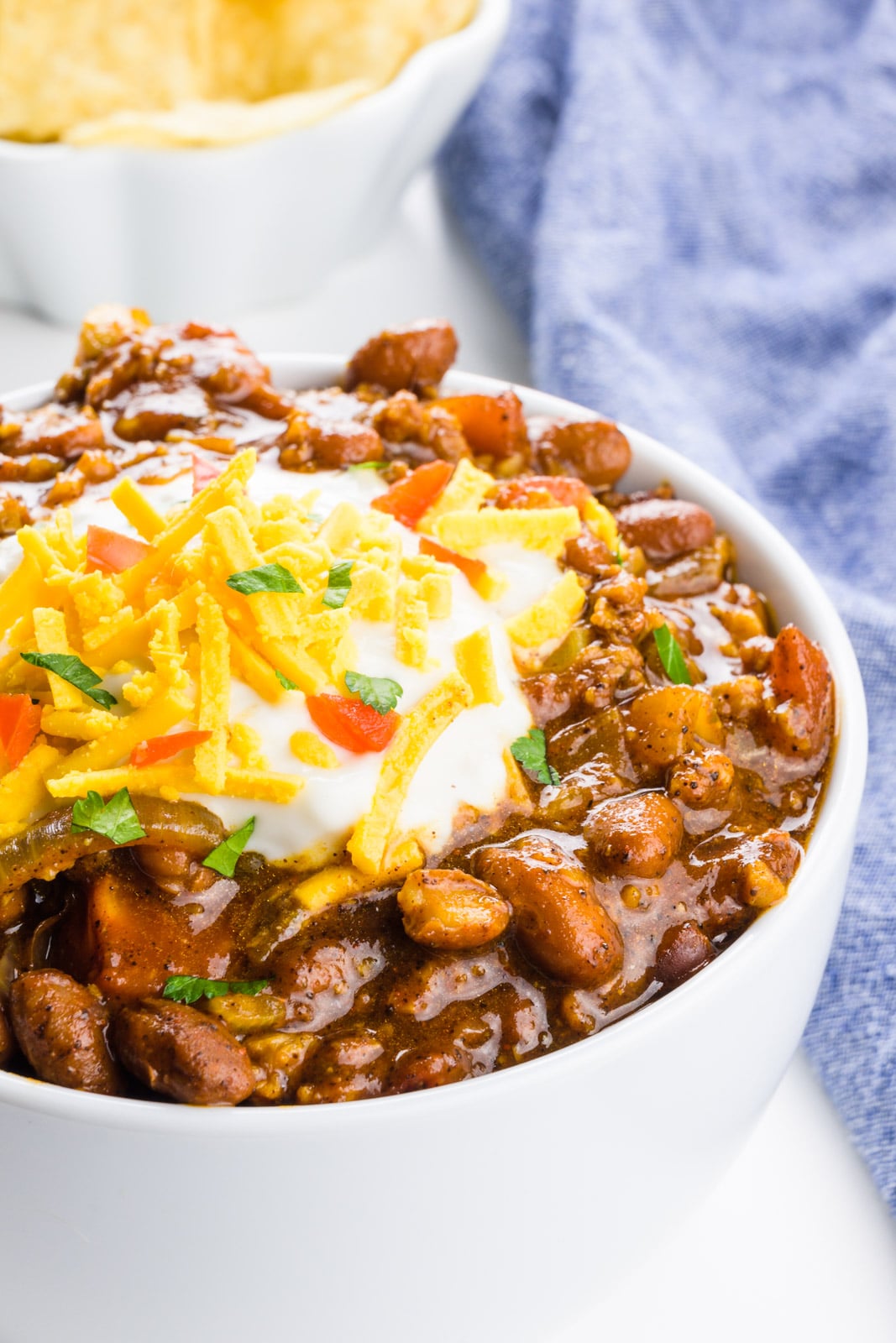 A bowl of vegan chili is topped with sour cream, and vegan cheddar shreds. A bowl of tortilla chips and a blue kitchen towel is behind it.