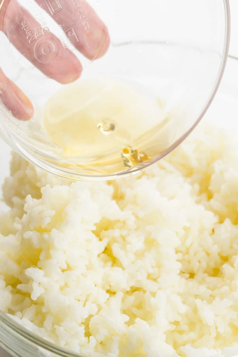 A hand holds a glass bowl, pouring liquid into another bowl full of white rice.