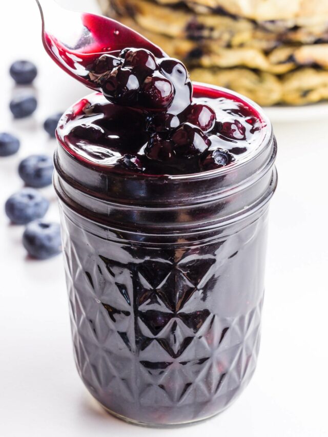 A spoon homers over a mason jar full of blueberry sauce. There are several waffles in the background.