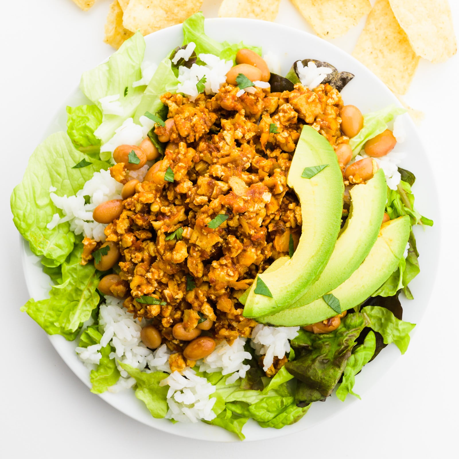 Looking down on a bowl of greens topped with rice, sofritas, and sliced avocados. There are tortilla chips by the plate too.