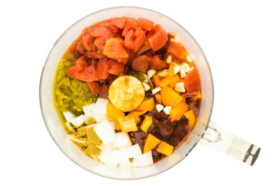 Looking down on a food processor full of ingredients, like onions, tomatoes, garlic, and more.
