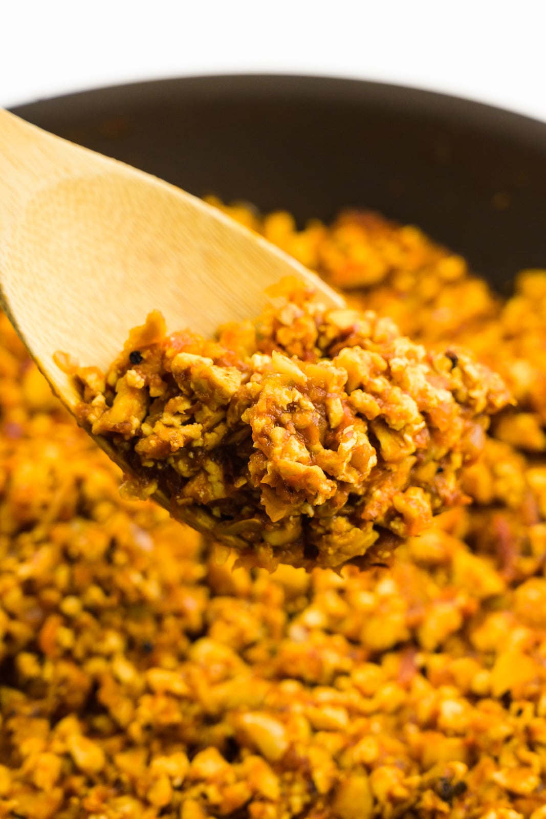 A wooden spoon holds a serving of sofritas made from tofu, hovering over a skillet full of more of it.