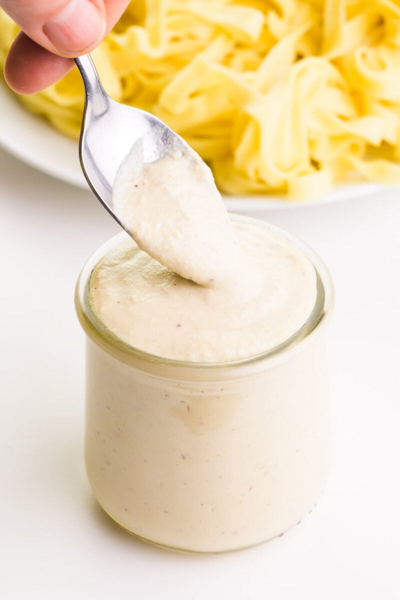 A spoon is scooping Alfredo Sauce out of a jar. There is a plate of fettuccine noodles in the background.