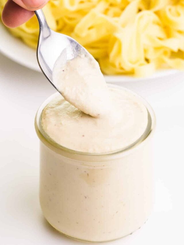 A spoon is scooping Alfredo Sauce out of a jar. There is a plate of fettuccine noodles in the background.