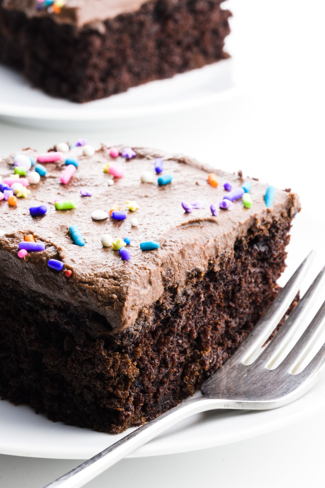 A fork sits beside a slice of wacky cake with chocolate frosting and sprinkles. Another slice of cake sits on a plate behind it.