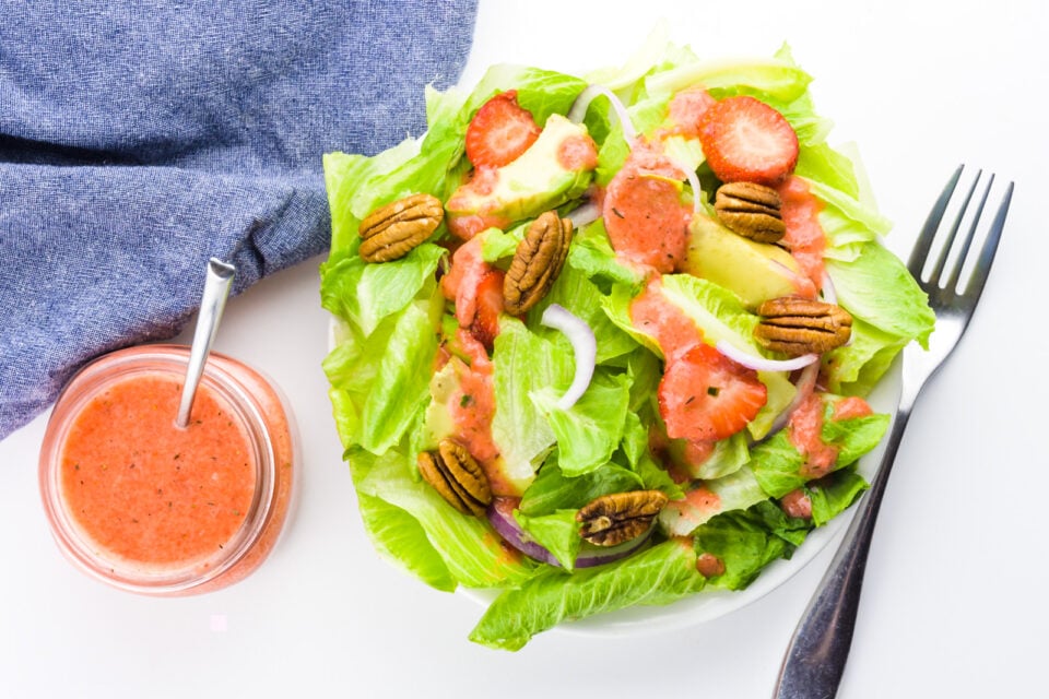 Looking down on a green salad with a fork beside it and a bowl of strawberry vinaigrette beside it too.