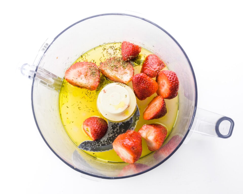 Chopped strawberries and other ingredients are in the bottom of a food processor bowl.