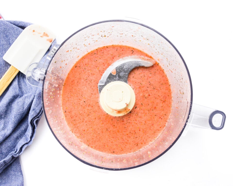 A pink mixture is in the bottom of a food processor bowl. A rubber spatula sits on a blue kitchen towel next to it.